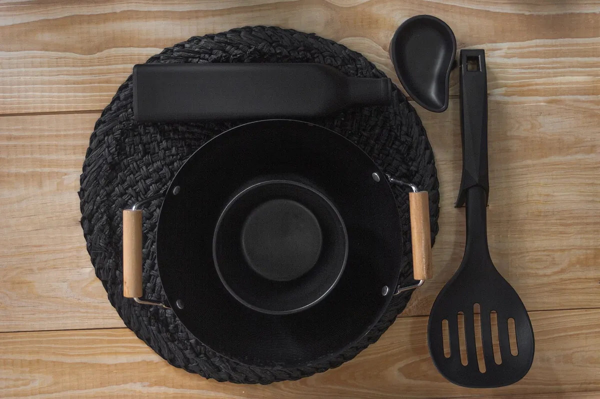 Black ceramic bowl, bottle, and utensils arranged on woven mat and wooden surface from above.