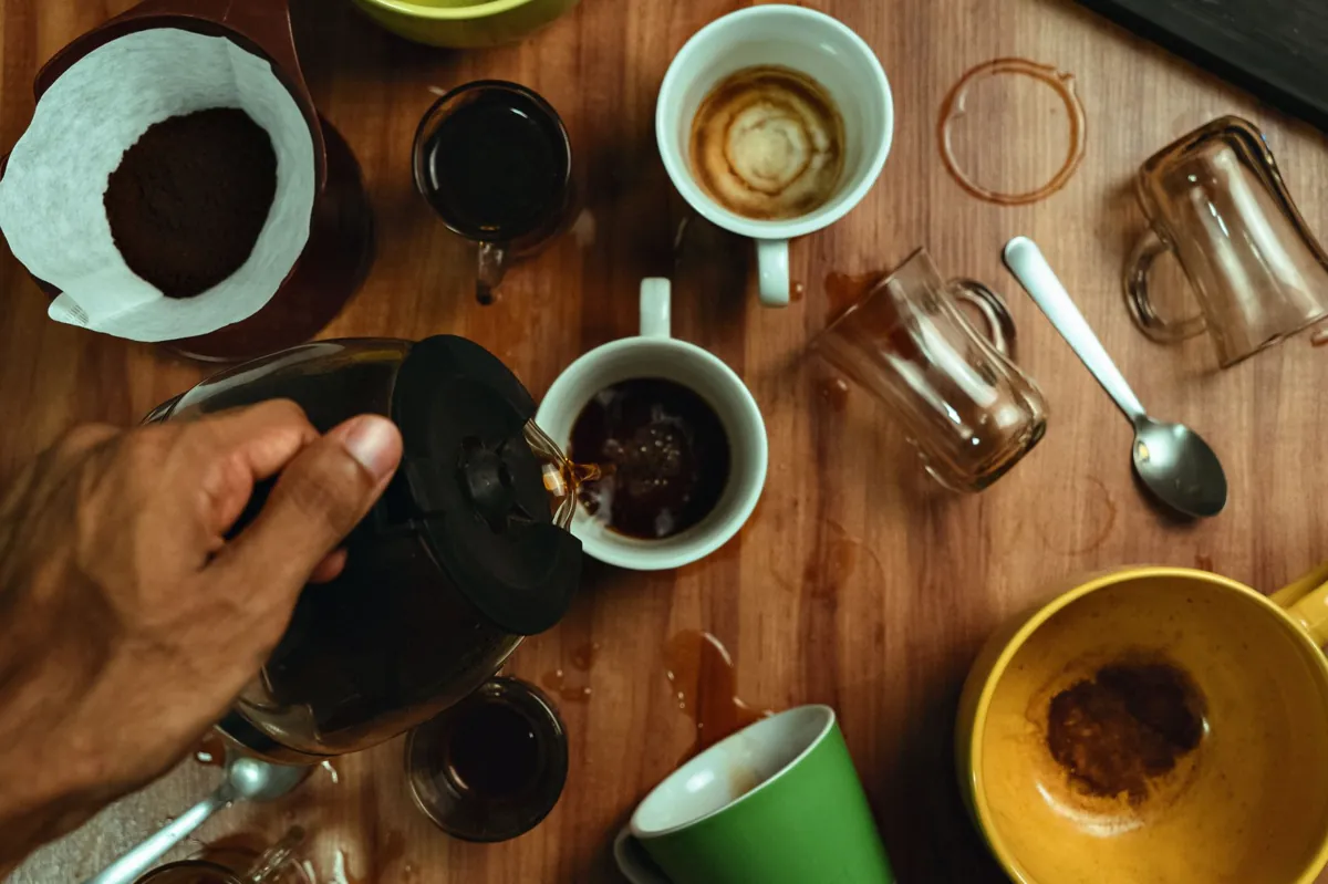 Top view of a hand pouring coffee into various cups on a messy wooden table.
