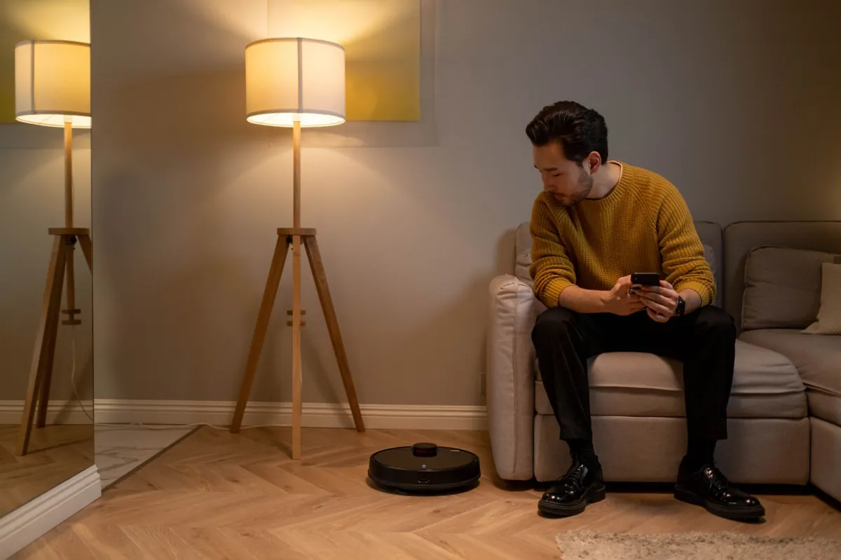 Adult man with a smartphone monitors robot vacuum in a modern living room.